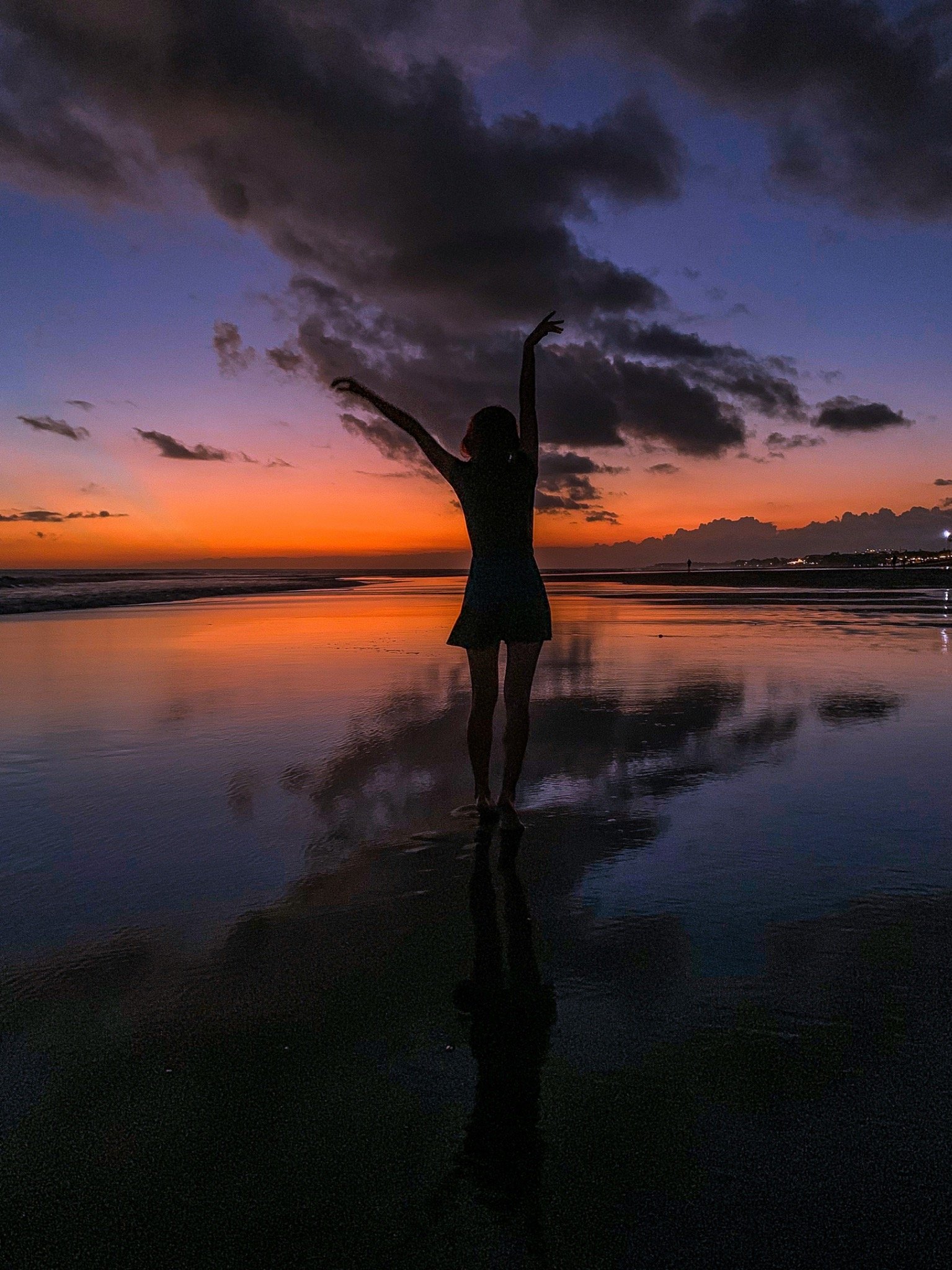 A woman standing at the water's edge with her arms raised against a twilight sky